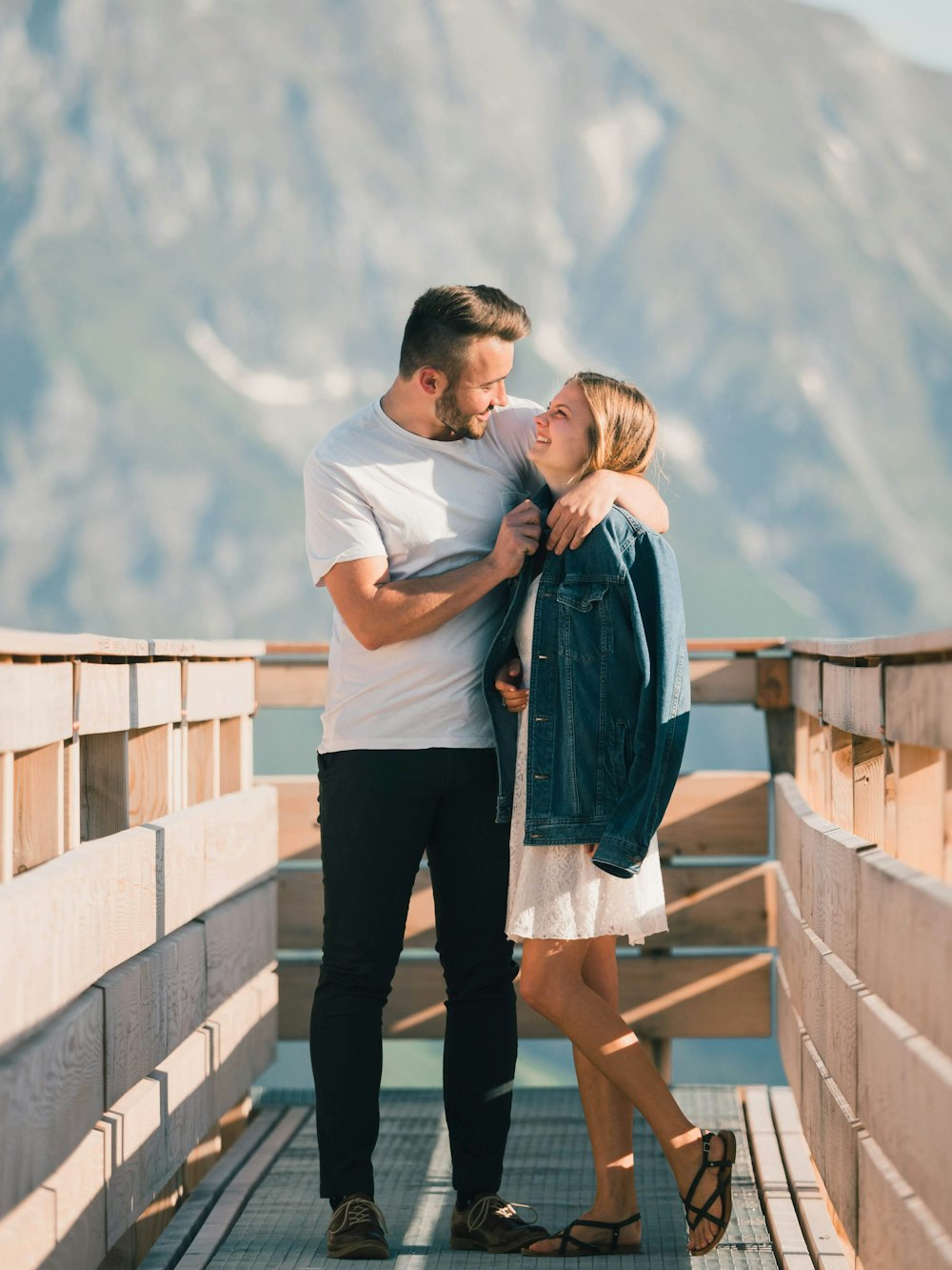 man in white t-shirt kissing woman in black pants on brown wooden fence during daytime