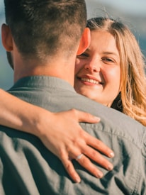 A woman with blonde hair smiles while embracing a man. The photo captures an intimate moment, focusing on the warmth and affection in her expression. She is wearing a ring on her hand, which rests on his back.