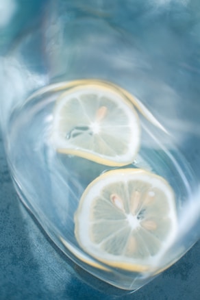 Two slices of lemon are submerged in a transparent liquid, likely water, within a container. The lemons are thinly sliced and positioned at slight angles, with their seeds visible. The background shows a textured blue surface that adds a cool tone to the composition.