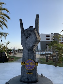 A large stone sculpture of a hand making the 'rock on' gesture is prominently displayed. It is located outdoors with palm trees and a hotel building in the background. The base of the sculpture features a logo and text indicating 'Hard Rock Hotel Tenerife.'
