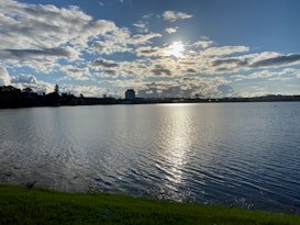 The image depicts a serene lake with calm water reflecting the sunlight. The sky is partly cloudy with a mixture of white fluffy clouds and clear blue patches. On the horizon, there are silhouettes of trees and a few buildings. The foreground shows a grassy edge along the lake.