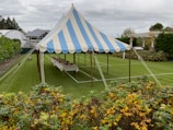 A blue and white striped tent is set up on a well-manicured lawn, surrounded by greenery and a garden with colorful plants in the foreground. Inside the tent, a long table is covered with a white cloth and arranged with various items. The sky is overcast, and there are buildings and a trellis visible in the background.