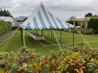 A blue and white striped tent is set up on a well-manicured lawn, surrounded by greenery and a garden with colorful plants in the foreground. Inside the tent, a long table is covered with a white cloth and arranged with various items. The sky is overcast, and there are buildings and a trellis visible in the background.