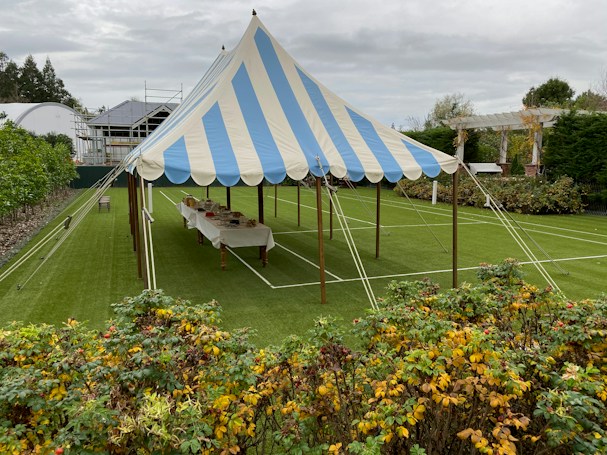 A blue and white striped tent is set up on a well-manicured lawn, surrounded by greenery and a garden with colorful plants in the foreground. Inside the tent, a long table is covered with a white cloth and arranged with various items. The sky is overcast, and there are buildings and a trellis visible in the background.