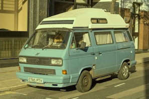 A vintage blue camper van is parked on the street beside a building. The van appears to be an older model with a high roof and windows covered with curtains. The exterior shows signs of wear, particularly noticeable around the wheel arches. The van has a white roof and is positioned partially on the sidewalk.