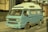 A vintage blue camper van is parked on the street beside a building. The van appears to be an older model with a high roof and windows covered with curtains. The exterior shows signs of wear, particularly noticeable around the wheel arches. The van has a white roof and is positioned partially on the sidewalk.