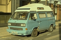 A vintage blue camper van is parked on the street beside a building. The van appears to be an older model with a high roof and windows covered with curtains. The exterior shows signs of wear, particularly noticeable around the wheel arches. The van has a white roof and is positioned partially on the sidewalk.