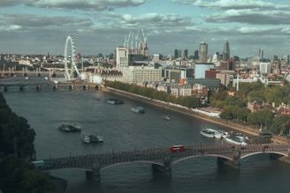 A view of a bustling cityscape featuring the London Eye, several bridges crossing the Thames River, and iconic skyscrapers in the background. Boats are seen navigating the river and a red double-decker bus is crossing one of the bridges.