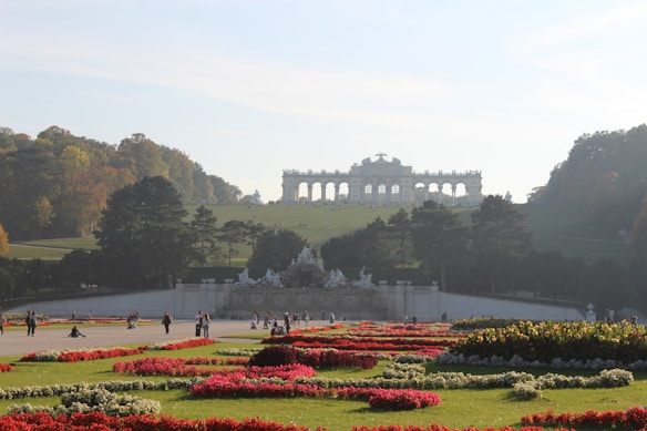 A beautifully landscaped garden with vibrant flower beds in various colors leading to a grand arch-like structure in the background. The scene is enveloped with lush greenery and scattered groups of people enjoying the serene environment.
