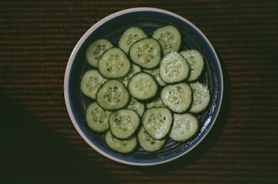 A ceramic bowl filled with fresh cucumber slices is placed on a dark, textured surface. The cucumbers are uniformly cut and arranged neatly, with seeds and flesh visible, reflecting a subtle sheen.