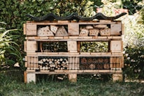 A garden insect hotel constructed from wooden pallets and filled with various natural materials like logs, bamboo, and pinecones. It is situated outdoors, surrounded by greenery and a wire fence. The structure is detailed with rows and compartments for different insects.