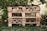 A garden insect hotel constructed from wooden pallets and filled with various natural materials like logs, bamboo, and pinecones. It is situated outdoors, surrounded by greenery and a wire fence. The structure is detailed with rows and compartments for different insects.