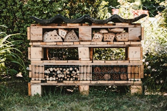 A garden insect hotel constructed from wooden pallets and filled with various natural materials like logs, bamboo, and pinecones. It is situated outdoors, surrounded by greenery and a wire fence. The structure is detailed with rows and compartments for different insects.