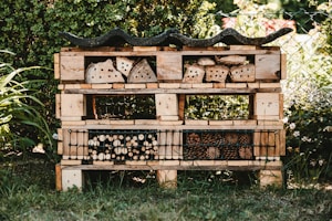 A garden insect hotel constructed from wooden pallets and filled with various natural materials like logs, bamboo, and pinecones. It is situated outdoors, surrounded by greenery and a wire fence. The structure is detailed with rows and compartments for different insects.