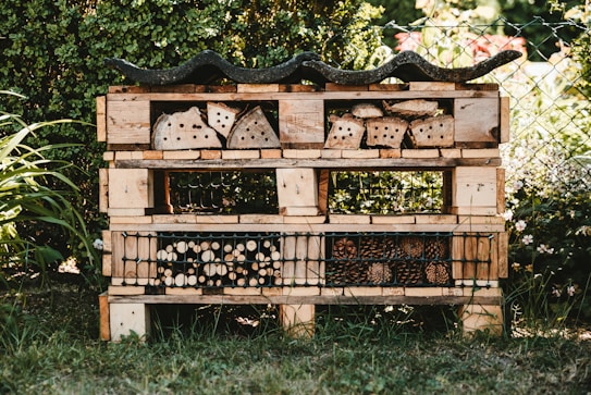 A garden insect hotel constructed from wooden pallets and filled with various natural materials like logs, bamboo, and pinecones. It is situated outdoors, surrounded by greenery and a wire fence. The structure is detailed with rows and compartments for different insects.