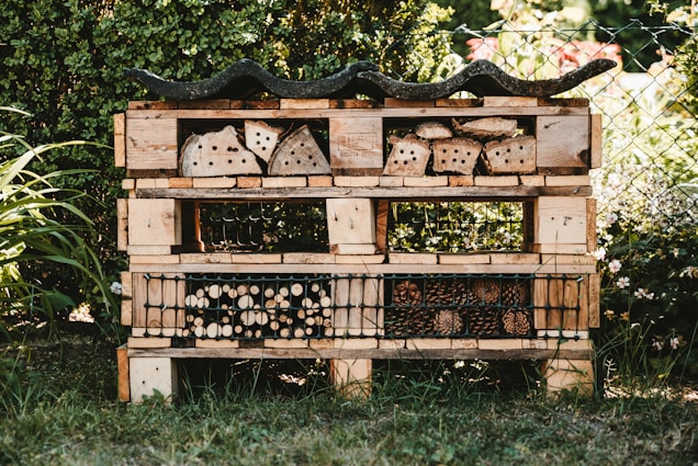 A garden insect hotel constructed from wooden pallets and filled with various natural materials like logs, bamboo, and pinecones. It is situated outdoors, surrounded by greenery and a wire fence. The structure is detailed with rows and compartments for different insects.