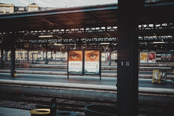 A train station platform with an overhanging metal roof and a large advertisement featuring a pair of eyes. The surroundings include multiple tracks, additional advertisements in the background, and architectural details of the station. The lighting suggests an early morning or late afternoon setting.