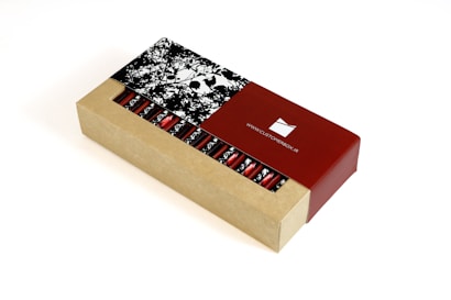 A rectangular cardboard box partially open to reveal a series of cylindrical red foil-wrapped items inside. The box features a contrasting design with a natural cardboard look and a glossy red section displaying a website URL. The lid has a monochrome floral pattern.