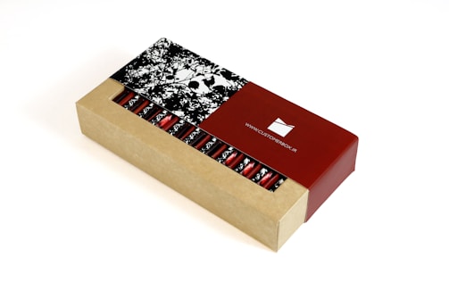 A rectangular cardboard box partially open to reveal a series of cylindrical red foil-wrapped items inside. The box features a contrasting design with a natural cardboard look and a glossy red section displaying a website URL. The lid has a monochrome floral pattern.