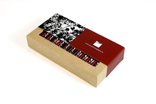 A rectangular cardboard box partially open to reveal a series of cylindrical red foil-wrapped items inside. The box features a contrasting design with a natural cardboard look and a glossy red section displaying a website URL. The lid has a monochrome floral pattern.