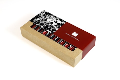 A rectangular cardboard box partially open to reveal a series of cylindrical red foil-wrapped items inside. The box features a contrasting design with a natural cardboard look and a glossy red section displaying a website URL. The lid has a monochrome floral pattern.