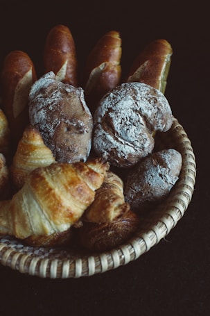 A basket filled with a variety of freshly baked breads including croissants and rustic loaves. The bread appears golden and crusty, dusted with flour, sitting against a dark background which accentuates their texture.