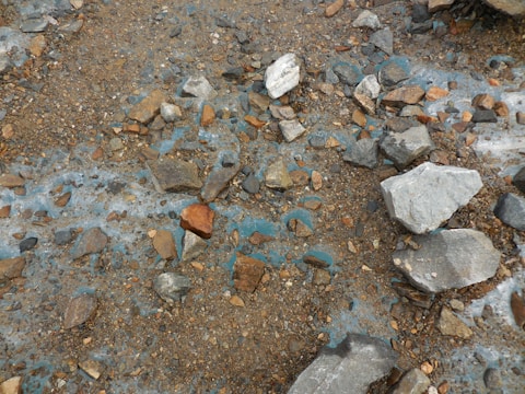 A rough, rocky terrain with a mix of small to medium-sized stones scattered across the surface. The ground appears to have patches of a bluish tint interspersed with brown earth and rocks. The rocks vary in color from gray to reddish-brown, giving a rugged and natural appearance.