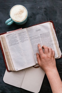 A close-up of a reader’s hand tracing lines in a helenimpress book, with a steaming cup of tea nearby and light shadows falling gently.
