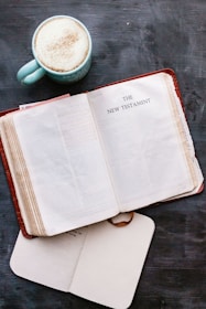 An open book displaying the title 'The New Testament' lies on a dark surface next to a cup of frothy coffee. The book has yellowed pages and seems well-used, suggesting age and frequent reading. A closed notebook or journal is partially underneath the book, adding a personal touch to the scene.
