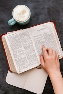 A hand is shown resting on an open Bible, specifically on a page that includes the book of 2 Corinthians. Next to the Bible, there is a light blue mug filled with frothy coffee on a dark surface, and an open notebook is mostly out of the frame beneath the Bible.