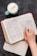 A hand is shown resting on an open Bible, specifically on a page that includes the book of 2 Corinthians. Next to the Bible, there is a light blue mug filled with frothy coffee on a dark surface, and an open notebook is mostly out of the frame beneath the Bible.