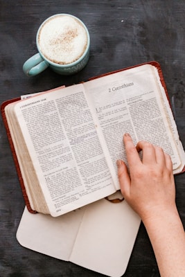 A hand is shown resting on an open Bible, specifically on a page that includes the book of 2 Corinthians. Next to the Bible, there is a light blue mug filled with frothy coffee on a dark surface, and an open notebook is mostly out of the frame beneath the Bible.