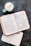 An open Bible displays text from 1 Corinthians, placed on a dark surface. A cup of frothy coffee in a light blue mug sits nearby alongside a closed notebook.