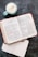 An open Bible displays text from 1 Corinthians, placed on a dark surface. A cup of frothy coffee in a light blue mug sits nearby alongside a closed notebook.
