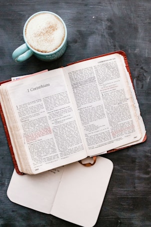 An open Bible displays text from 1 Corinthians, placed on a dark surface. A cup of frothy coffee in a light blue mug sits nearby alongside a closed notebook.