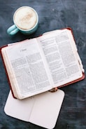 An open book with a brown leather cover displaying a passage from 1 Peter, accompanied by a cup of coffee in a light blue mug on a dark blue surface. An additional open white notebook lies beneath the book.