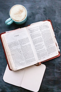 An open book with a brown leather cover displaying a passage from 1 Peter, accompanied by a cup of coffee in a light blue mug on a dark blue surface. An additional open white notebook lies beneath the book.