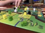A close-up of a board game featuring colorful wooden pieces in various shapes and colors, placed on a detailed game board. A person’s hand is interacting with the game, indicating active play. The board has a vibrant design with green, yellow, and brown areas.