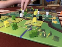 A close-up of a board game featuring colorful wooden pieces in various shapes and colors, placed on a detailed game board. A person’s hand is interacting with the game, indicating active play. The board has a vibrant design with green, yellow, and brown areas.