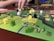 A close-up of a board game featuring colorful wooden pieces in various shapes and colors, placed on a detailed game board. A person’s hand is interacting with the game, indicating active play. The board has a vibrant design with green, yellow, and brown areas.