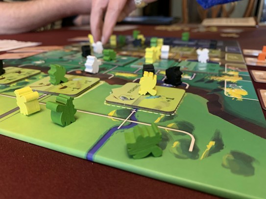 A close-up of a board game featuring colorful wooden pieces in various shapes and colors, placed on a detailed game board. A person&rsquo;s hand is interacting with the game, indicating active play. The board has a vibrant design with green, yellow, and brown areas.