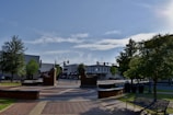 A small town square with brick paved walkways and a circular seating area in the foreground. Mature trees provide shade, and street lamps line the square. In the background, small shops and buildings with signage are visible under a clear blue sky.