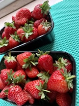 Two black plastic containers filled with freshly picked strawberries are placed on a turquoise textured mat. The strawberries are red and ripe with green leaves still attached, suggesting they are fresh and ready to eat.