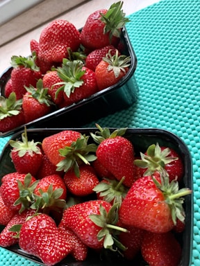 Two black plastic containers filled with freshly picked strawberries are placed on a turquoise textured mat. The strawberries are red and ripe with green leaves still attached, suggesting they are fresh and ready to eat.