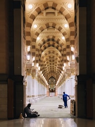 A spacious and ornately decorated interior hallway is depicted with repeating arched ceilings adorned with geometric patterns and warm lighting. There are two individuals visible; one is sitting on the floor near the entrance, and the other appears to be a worker in blue uniform operating a cleaning machine.