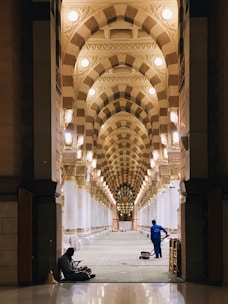 A spacious and ornately decorated interior hallway is depicted with repeating arched ceilings adorned with geometric patterns and warm lighting. There are two individuals visible; one is sitting on the floor near the entrance, and the other appears to be a worker in blue uniform operating a cleaning machine.