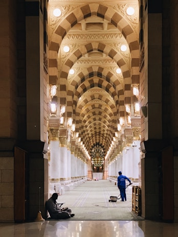 A spacious and ornately decorated interior hallway is depicted with repeating arched ceilings adorned with geometric patterns and warm lighting. There are two individuals visible; one is sitting on the floor near the entrance, and the other appears to be a worker in blue uniform operating a cleaning machine.
