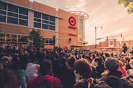 A large crowd of people gather peacefully in front of a retail store under a partially cloudy sky. The storefront features a prominent logo and signage. Participants appear engaged and focused, with some holding signs. The scene suggests a demonstration or protest, with the event taking place in an urban environment.