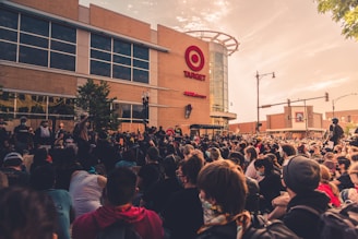 A large crowd of people gather peacefully in front of a retail store under a partially cloudy sky. The storefront features a prominent logo and signage. Participants appear engaged and focused, with some holding signs. The scene suggests a demonstration or protest, with the event taking place in an urban environment.