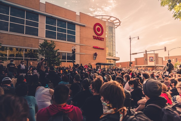 A large crowd of people gather peacefully in front of a retail store under a partially cloudy sky. The storefront features a prominent logo and signage. Participants appear engaged and focused, with some holding signs. The scene suggests a demonstration or protest, with the event taking place in an urban environment.